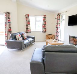 A living room with two black sofas a wooden coffee table a wall-mounted TV and windows with patterned curtains at Callow Lodge 22 in Beaconsfield Holiday Park
