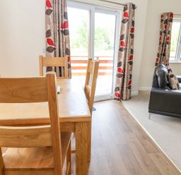 A dining area with wooden chairs and table next to a sliding glass door with leaf patterned curtains beside a living room with a black sofa and matching curtains at Callow Lodge 22 in Beaconsfield Holiday Park