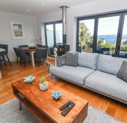 A living room with a gray sofa and wooden coffee table near dining table and large windows overlooking the sea at Seafield in Carbis Bay near St Ives