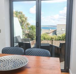 A dining area with a wooden table and blue chairs next to glass doors opening to a patio with wicker furniture and sea view at Seafield in Carbis Bay near St Ives