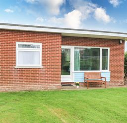 A brick building with a white door and windows a wooden bench and a potted plant on grass at 20 Siesta Mar in Mundesley
