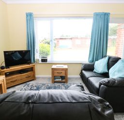 A living room with black leather sofas and blue cushions a wooden TV stand with a television a small wooden side table and blue curtains at 20 Siesta Mar in Mundesley