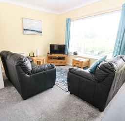A living room with two black leather sofas a wooden TV stand and side tables and a large window with blue curtains at 20 Siesta Mar in Mundesley
