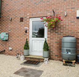 A brick house exterior with a white door flanked by two potted plants a hanging basket with pink flowers and a rain barrel at The Annex @ Garden House in Prudhoe near Newcastle