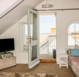A room with a patchwork sofa and TV near a door opening to a balcony overlooking a beach at Spiral View in Perranporth
