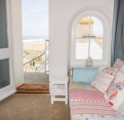 A seating area with a patterned sofa and cushions next to a window and an open door leading to a balcony overlooking the beach at Spiral View in Perranporth