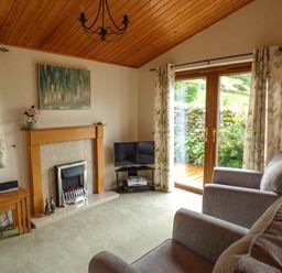A living room with armchairs a fireplace a television and glass doors leading outside at South Lodge in Kendal