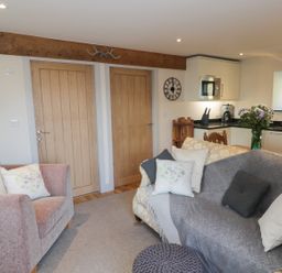 A living area with a pink upholstered armchair, two wooden doors, a gray sofa with cushions, a kitchen with white cabinets and appliances, and a wooden dining table with flowers at The Granary Loft in Bardsea