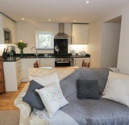 A living room with a sofa and cushions open to a kitchen with white cabinets and appliances at The Granary Loft in Bardsea