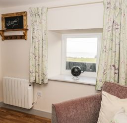 A window with floral curtains a small globe on the sill a wall-mounted radiator a brown sofa with cushions and a wooden framed welcome sign on the wall at The Granary Loft in Bardsea