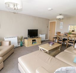 A living room with beige sofas a wooden coffee table a television on a wooden stand and a dining table with chairs at Harbour View in Cemaes Bay