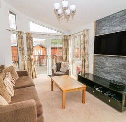 A living room with a brown sofa and armchair a wooden coffee table a wall-mounted TV and large windows with curtains at Coniston Number 9 in White Cross Bay