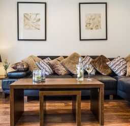 A living room with a black leather sofa with multiple cushions a wooden coffee table with a bottle and glass of white wine a small side table with a lamp and a vase with flowers and two framed artworks on the wall at Tus Nua Cottage in Carhampton