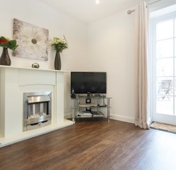 A room with a white fireplace decorated with two vases and a painting a TV on a glass stand and a door leading outside at Tus Nua Cottage in Carhampton