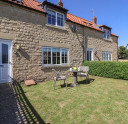 A stone house with a red tiled roof a white door and windows a small patio table with two chairs on a grassy lawn at 8 Pottergate Mews Helmsley