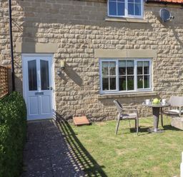 An outdoor garden area with two chairs and a table with a teapot and mugs in front of a stone house with a white door and windows at 8 Pottergate Mews in Helmsley