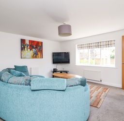 A living room with a curved blue sofa a wall mounted television a wooden door and a window with striped blinds at 8 Pottergate Mews Helmsley