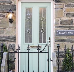 A green front door with decorative glass panels and a metal handle set in a stone wall with a light above and a black metal fence in front at Llwyn Onn Dolwyddelan