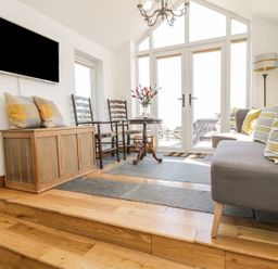 A sitting area with a gray sofa with striped cushions wooden chairs a small round table with flowers and a wall-mounted television at Marsh Villa in Silloth