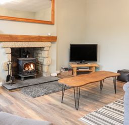 A living room with a stone fireplace a wood burning stove a wooden coffee table a TV on a wooden stand and a striped rug at Victoria Cottage in Norham