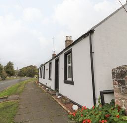 A white cottage with black window frames and a paved sidewalk in front near a road with grass and trees at Victoria Cottage in Norham