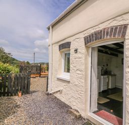 Exterior view of a cottage with a doorway to a kitchen and a small fenced garden area with chairs at Taliharris Cottage in Llandysul