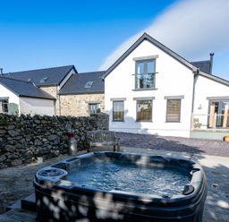 A hot tub near a stone wall and a house with multiple windows and doors at Sgubor in Pentraeth