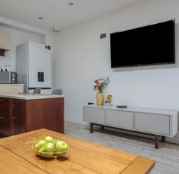 A kitchen and living area with a wooden table holding green apples a white cabinet with a vase and a TV mounted on the wall at The Studio @ The Old Post Office in Cemaes Bay