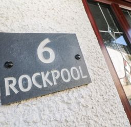 A black house number plaque with number 6 and text Rockpool on a rough white wall near a door with decorative glass panels at Rockpool in Harlyn Bay near St Merryn