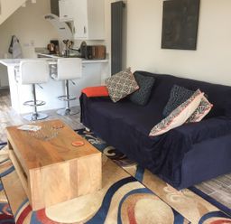 A living room with a sofa covered with a dark cloth and patterned cushions a wooden coffee table two white bar stools and a kitchen counter at Squirrels Drey Little Dunham near Litcham
