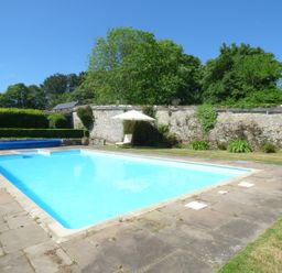 An outdoor swimming pool surrounded by stone paving and grass with a stone wall and trees at Parlour Cottage in Marazion
