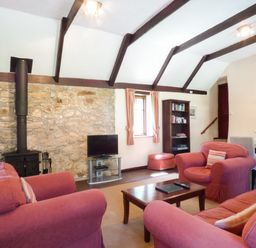A living room with red armchairs, a wooden coffee table, a wall-mounted stove, a stone feature wall, a television, a bookshelf, and a dining area at Parlour Cottage in Marazion