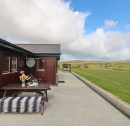 An outdoor patio area with a wooden picnic table with a flower vase and cups and a bench with a blanket beside a brown wooden building with fields and blue sky in the background at Chywolow Lodge in Penzance