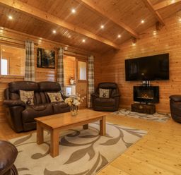 A wooden living room with leather sofas a wooden coffee table a rug a wall mounted TV and an electric fireplace at Chywolow Lodge in Penzance