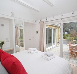 A bedroom with a bed and red pillows next to glass doors opening to a patio with a table and chairs at Cot Valley Cottage in St Just