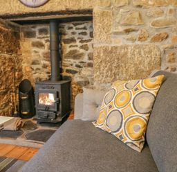 A living room with a wood burning stove set in a stone fireplace and a gray sofa with a patterned cushion at Pippin in Lelant