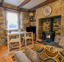 A room with a dining table and chairs next to a window a stone fireplace with a wood stove a wall clock above the fireplace and a sofa with patterned cushions at Pippin in Lelant