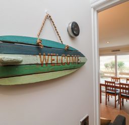 A wooden welcome sign hanging on a wall next to a thermostat with a dining area and sliding doors overlooking outdoor plants at Ocean Blue in Porthtowan
