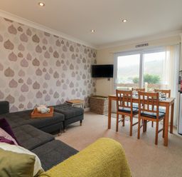 A living room with a grey sofa purple cushions a wooden dining table and chairs a wall mounted tv and sliding glass doors at Ocean Blue in Porthtowan