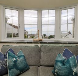 A sofa with blue and green cushions under a bay window with a small sailboat decoration at Bayview in Watergate Bay