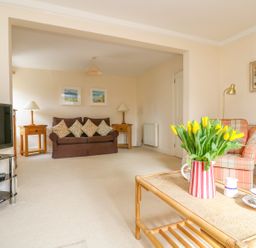 A living room with a brown sofa with cushions two wooden side tables with lamps a plaid armchair a coffee table with yellow flowers and tea set a tv on a stand and paintings on the walls at Goshen Farm in St Agnes