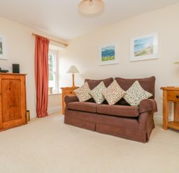 A living room with a brown sofa with patterned cushions wooden side tables with lamps wooden cabinet and framed pictures on the wall at Goshen Farm in St Agnes