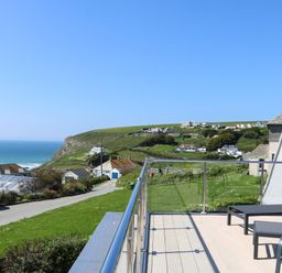A balcony with outdoor chairs and a table overlooking a coastal village and the ocean at Trelawns in Mawgan Porth