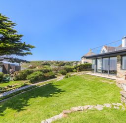 A grassy garden with shrubs and a stone path next to a house with large windows at Trelawns in Mawgan Porth