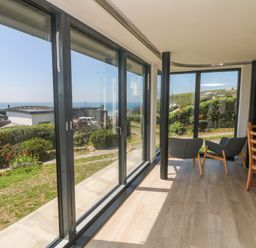 A room with large glass windows two gray chairs a wooden table and chairs overlooking a garden and ocean view at Trelawns in Mawgan Porth
