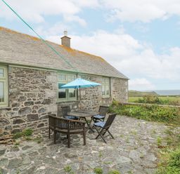 An outdoor patio with a wooden table and chairs with a blue umbrella next to a stone house with green windows at Parc An Castle Cottage in Lizard