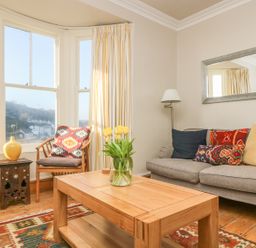 A living room with a gray sofa with colorful cushions a wooden coffee table with a vase of yellow tulips a wooden chair with a patterned cushion a small wooden side table with a yellow vase a floor lamp a wall mirror and large windows with curtains at Haven in Porthleven
