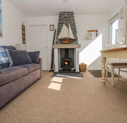 A living room with a gray sofa a stone fireplace with a model boat and a wooden dining table at Cockle Island Cottage in Porthallow