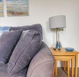 A sofa with cushions and a blue check blanket next to a wooden nesting table with a lamp and bowl at Cockle Island Cottage in Porthallow
