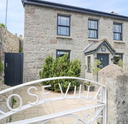 Stone house with black framed windows and a small porch behind a white metal gate with rose villa on the gate at Rose Villa in Newlyn